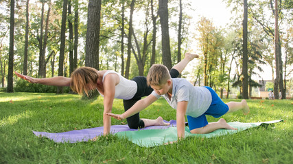 Photo of mother with her teenage boy son practising yoga asana on grass at park. Family doing fitness and sports outtodr at forest