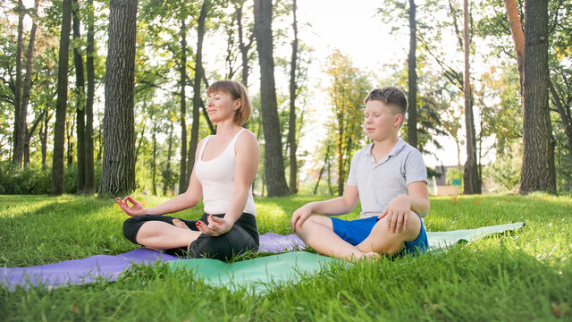 Photo of middle aged woman teaching her student in yoga class at park. Woamn with teenage boy practising fitness, meditation and yoga on grass at forest