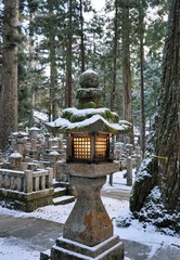Old stone lantern covered in moss and snow, light inside it, in the Mount Koya Buddhist cemetery, Japan 