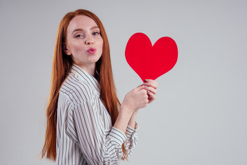 Happy redhead girl businesswoman in a striped shirt model sending air kiss with decorative hearts Valentines Day gift on white background studio