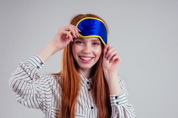 Good Morning. Smiling redhead young business woman in a striped shirt and blue yellow sleep mask white background studio