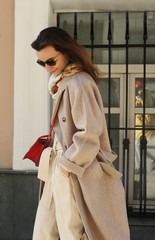 Portrait of a beautiful trendy fahsionista brunette with red lipstick in a beige coat with sunglasses. Posh stylish girl in Moscow. Black friday idea. Set sail champagne trendy color