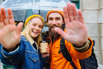 Hipster couple traveler under transparent umbrella in bright casual clothes saluting you, saying...