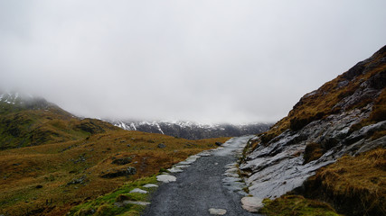 Stone path along an impactful view, surrounded by saturated grass, beautiful mountains and a foggy sky – captured during a hike at Snowdon in winter (Snowdonia National Park, Wales, United Kingdom)