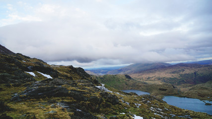 Fototapeta na wymiar Stunning mountain view from the top seeing a lake, rugged valley and moody skies – captured during a hike at Snowdon in winter (Snowdonia National Park, Wales, United Kingdom)
