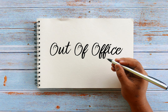 Top view of hand holding pen writing Out Of Office on notebook on wooden background.