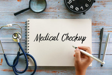 Top view of stethoscope,magnifying glass,clock,pen and hand holding pen writing Medical Checkup on notebook on wooden background.