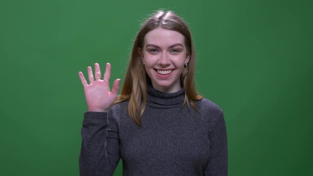 Pretty blonde model waves hand to camera being happy and friendly isolated on green chromakey background.