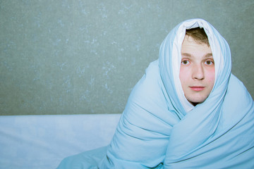 young guy sitting on a bed wrapped in a blue blanket