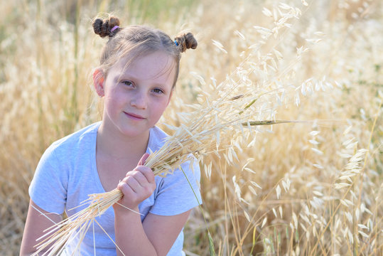Beautiful girl holding spikes of wheat and ears of oats. Cute child sitting on the gold autumn field ready for harvest. - Powered by Adobe