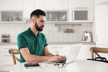 Handsome young man working with laptop at table in kitchen