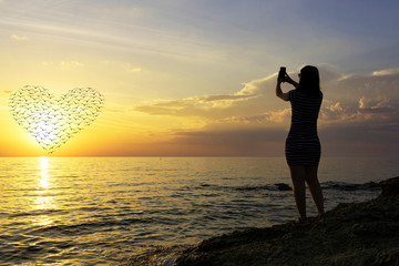 silhouette of a young girl. Photographs a beautiful sunset on the phone. heart of the silhouettes of birds in the sun.