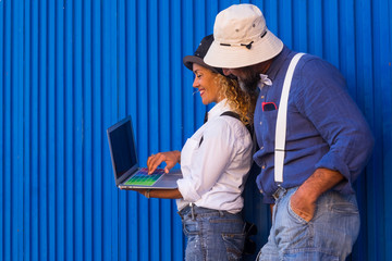 Happy caucasian couple man and woman dressed in a fun way with colored suspenders and hat looking at laptop. Lean against a blue shutter in the street. Sunlight
