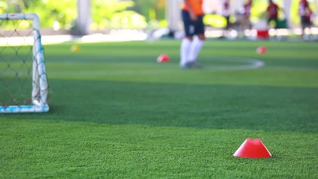 red marker cone on green artificial turf with blurry soccer team training and mini goal, blurry boy soccer player jogging between marker cones and control ball with soccer equipment in soccer academy.