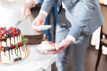 Obraz na płótnie Canvas Bride and groom cut beautiful rustic wedding cake on wedding banquet. The cake is decorated with fresh berry, strawberry, raspberry and chocolate. Fashionable luxury stylish cake