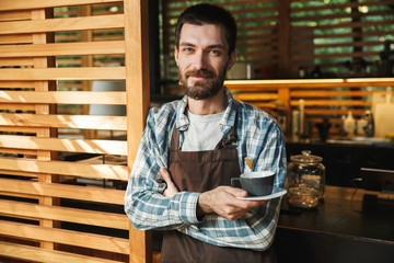 Portrait of kind barista man holding cup of coffee while working in street cafe or coffeehouse outdoor
