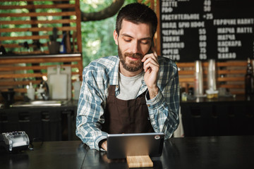 Portrait of joyous barista man using tablet computer while working in street cafe or coffeehouse outdoor