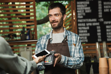 Portrait of friendly barista man holding consumer terminal for paypass payment while working in street cafe or coffeehouse outdoor