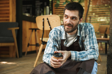 Photo of brunette waiter man sitting on wooden floor while working in cafe or coffeehouse outdoor