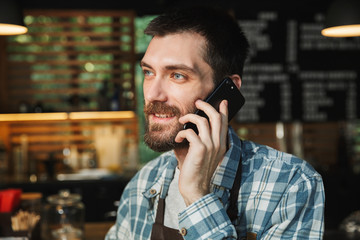 Portrait of happy barista guy talking on cellphone in street cafe or coffeehouse outdoor