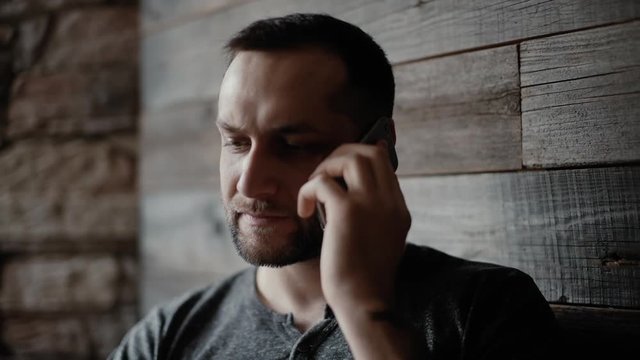 Close-up of brutal man with bristle and tattooed hands sits at a table in a cafe and speak by phone on the background of a beautiful wooden plank wall.