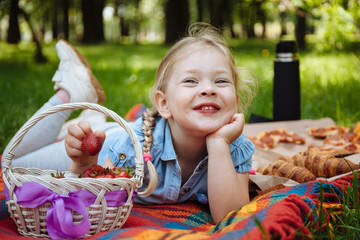 Cheerful little girl eating strawberries at picnic in park, sitting on green grass and smiling. Summer holidays and recreation.