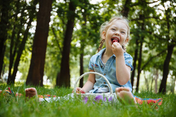 Cheerful little girl eating strawberries at picnic in park, sitting on green grass and smiling. Summer holidays and recreation.