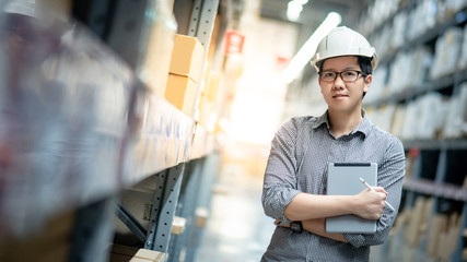 Young Asian man worker wearing safety helmet and eyeglasses doing stocktaking of product in cardboard box on shelves in warehouse by using digital tablet and pen. Physical inventory count concept