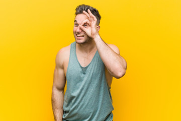 Young fitness man against a yellow background excited keeping ok gesture on eye.