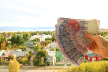 Female's Hand Holding Bunch of Argentinian 100 Pesos Bills(back) with Blurry EL Calafate Town in Background 