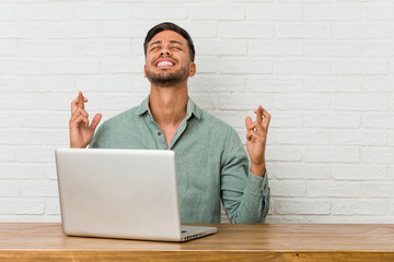 Young filipino man sitting working with his laptop crossing fingers for having luck
