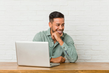 Young filipino man sitting working with his laptop keeps hands under chin, is looking happily aside.