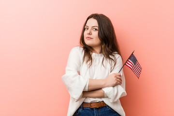 Young curvy plus size woman holding a united states flag smiling confident with crossed arms.