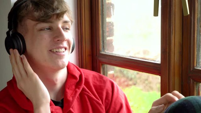 Happy smiling male teenager young man teen wearing a red hoodie, sitting by a window listening to music on wireless bluetooth headphones and cell phone