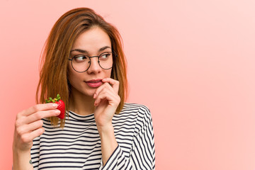 Young woman holding a strawberry relaxed thinking about something looking at a copy space.