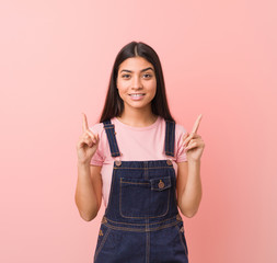 Young pretty arab woman wearing a jeans dungaree indicates with both fore fingers up showing a blank space.