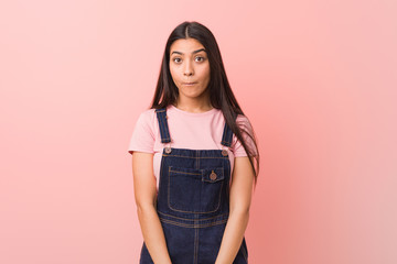 Young pretty arab woman wearing a jeans dungaree shrugs shoulders and open eyes confused.