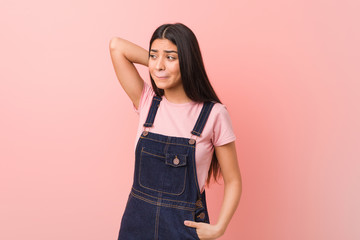 Young pretty arab woman wearing a jeans dungaree touching back of head, thinking and making a choice.