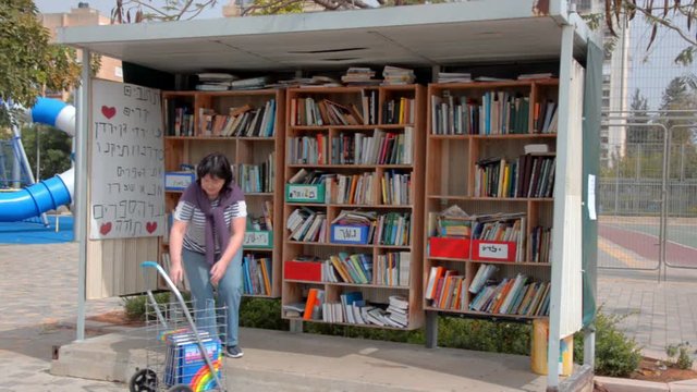 Mature woman brought some books in the 4-wheel folding shopping cart. She fills the street library at Hoshen street in Ness Ziona, Israel