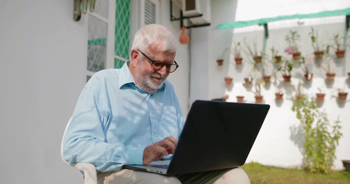 elderly serious man uses a laptop sitting on a chair in his backyard