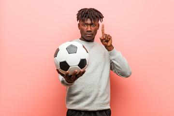 Young fitness black man holding a soccer ball showing number one with finger.