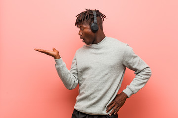 Young rasta black man listening to music with headphones impressed holding copy space on palm.