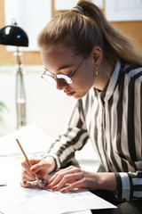 Naklejka na ściany i meble Photo of focused european woman architect wearing glasses designing draft and sitting at workplace