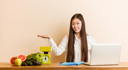 Obraz na płótnie Canvas Young nutritionist chinese woman working with her laptop showing a copy space on a palm and holding another hand on waist.