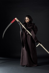full length view of pensive woman in death costume holding scythe on black