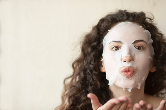 Beautiful young woman is applying a cosmetic tissue mask on a face on a white background 