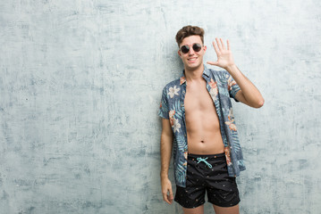 Fototapeta na wymiar Young caucasian man wearing a swimsuit smiling cheerful showing number five with fingers.