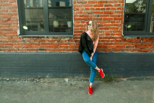 blonde girl with curly hair in black glasses, pink turtleneck, in a black leather rocker jacket on the background of a red brick wall, with a place for the inscription on the left