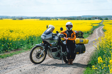 Fototapeta na wymiar Girl in a protection outfit and glasses with touristic motorcycle. Rape yellow flowers field on background. Adventure trail tour, enduro and off road, summer day. extreme vacation concept