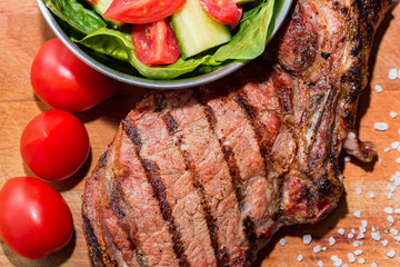 Close up freshly grilled bone steak on wood and fresh salad in bowl